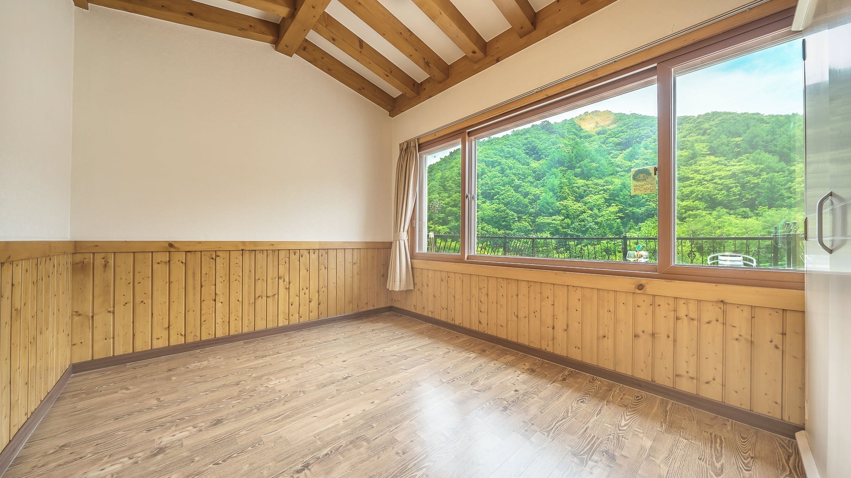 A room with wooden paneling, a large window with a view of a forest, and a wooden floor.