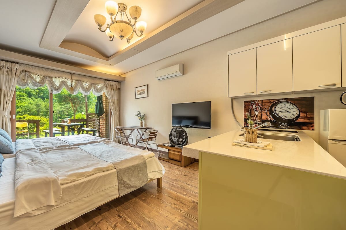 Quarto com uma cama grande, uma escrivaninha e uma televisão. Vê-se vegetação pela janela.