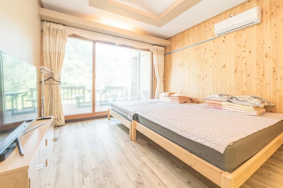 Quarto com piso de madeira, uma cama com colcha, televisão e janela com cortinas.