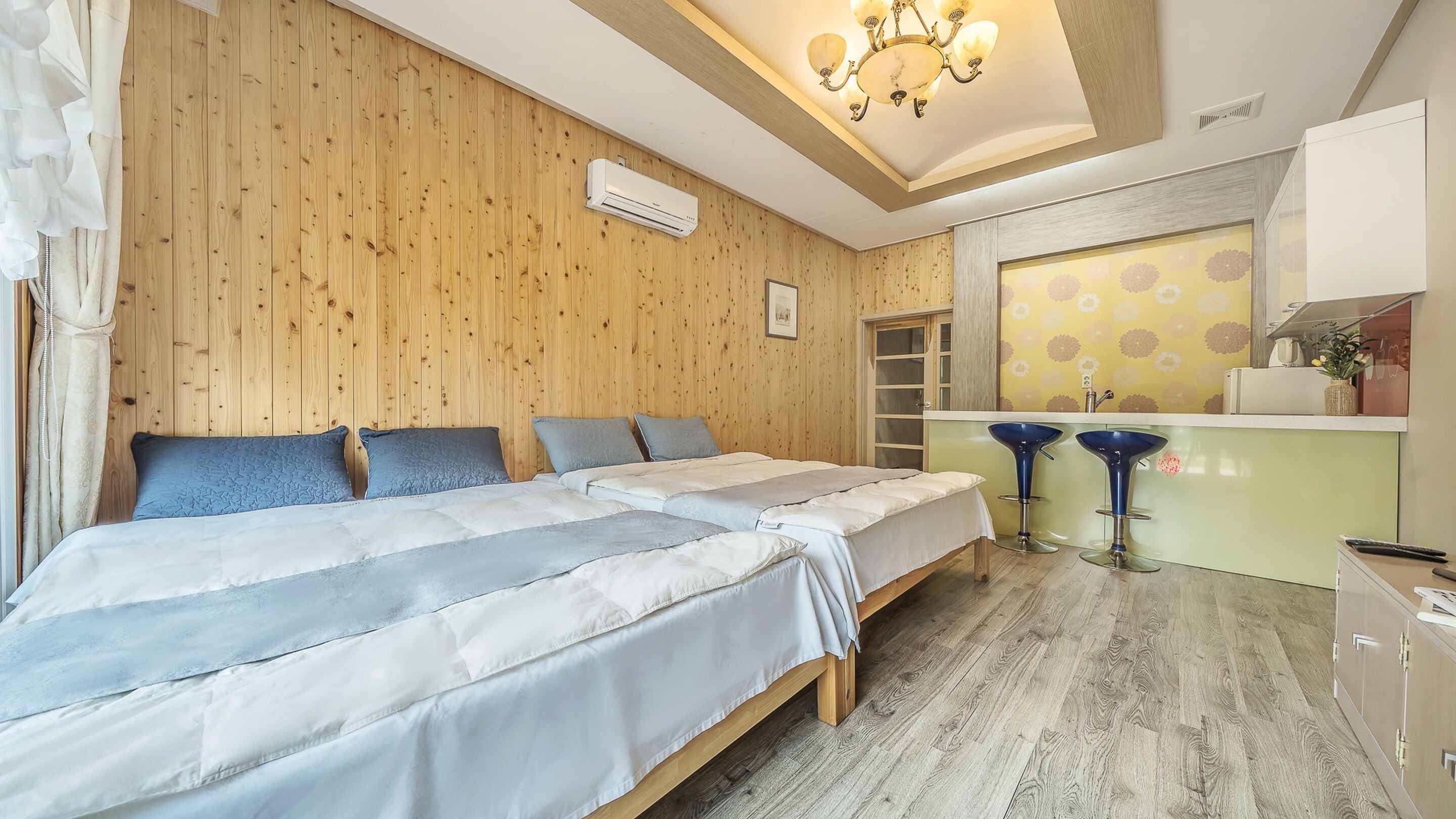 A room with a wooden wall, a bed with white and blue bedding, a small kitchen area with a sink and bar stools, and a chandelier.