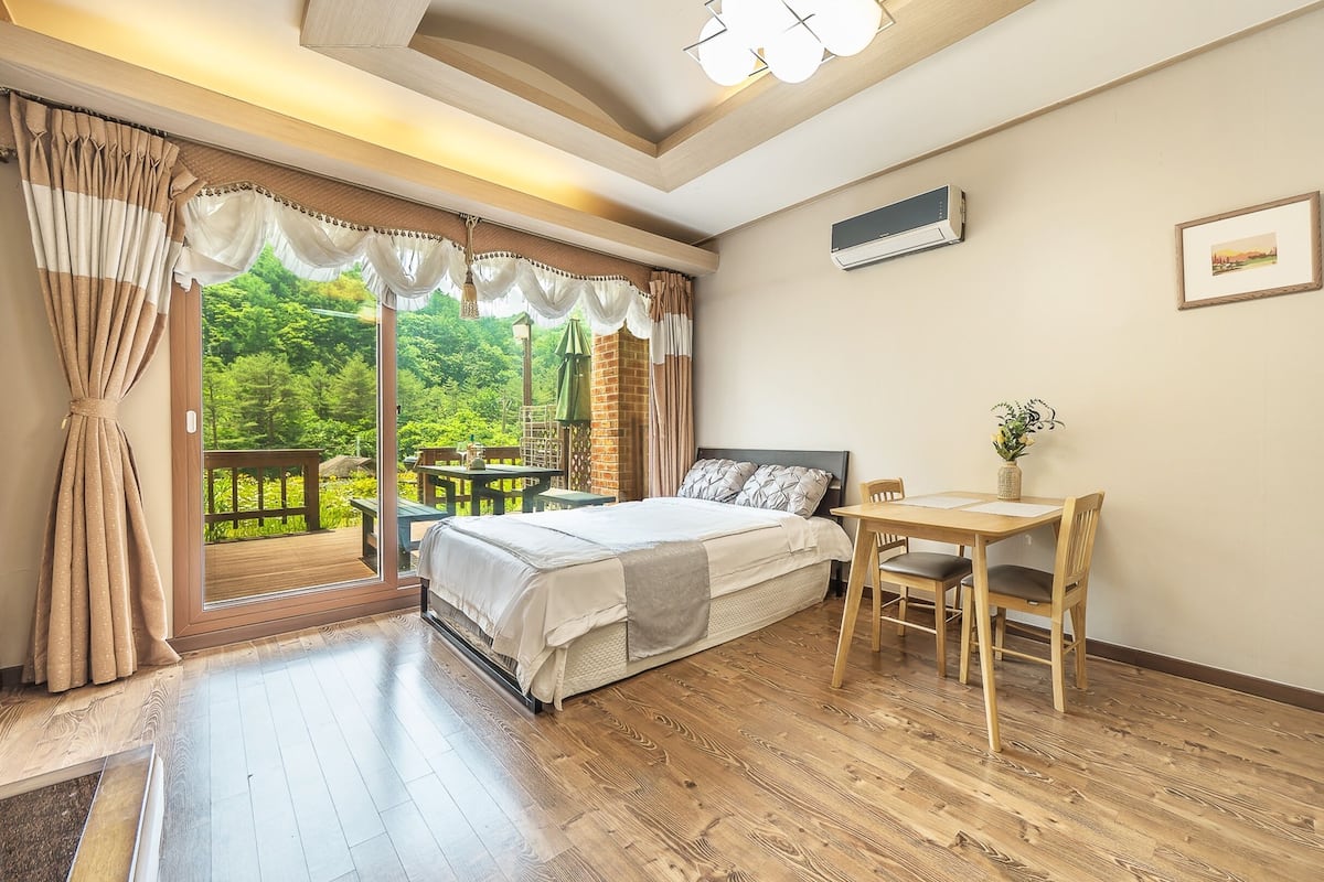 Quarto com uma cama, uma mesa de madeira e uma cadeira. Há uma janela com cortinas e vista para o verde lá fora.