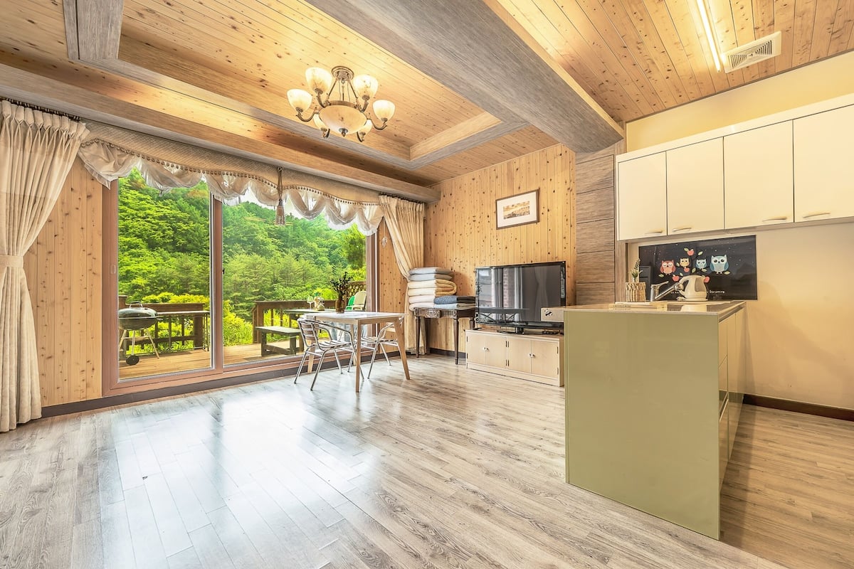Um quarto amplo com piso de madeira, sala de jantar com mesa e cadeiras, cozinha com TV e armários, e uma grande porta de vidro deslizante que dá para uma varanda com vegetação.