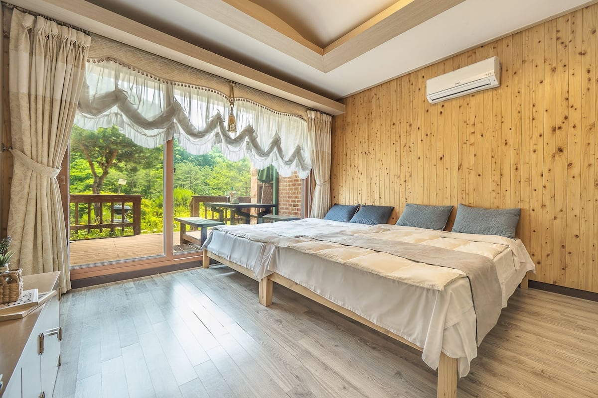 Quarto com cama grande, paredes de madeira, janela com cortinas e vista para o verde lá fora.
