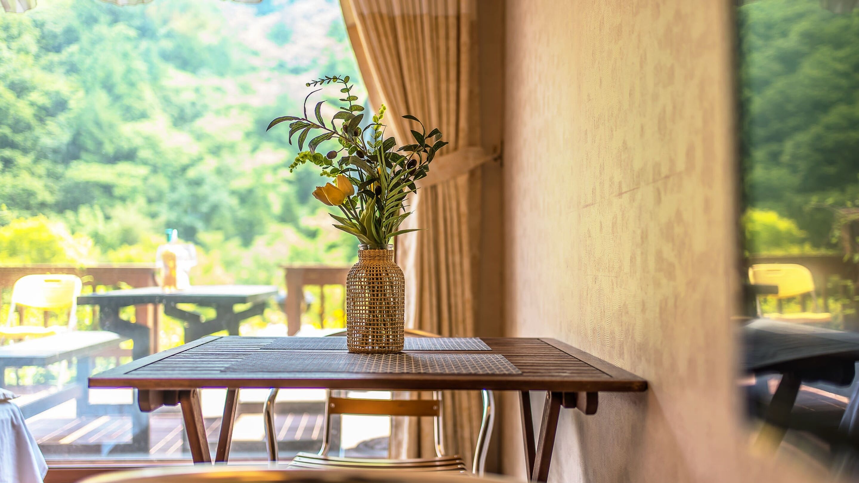 A dining table with a woven vase containing flowers, set against a backdrop of greenery.