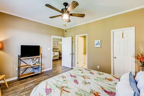 A bedroom with a bed, a ceiling fan, a TV on a stand, and a door leading to another room.