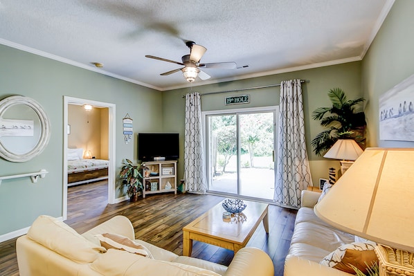 A living room with a sofa, a coffee table, a ceiling fan, a television, and a sliding glass door leading to a balcony.