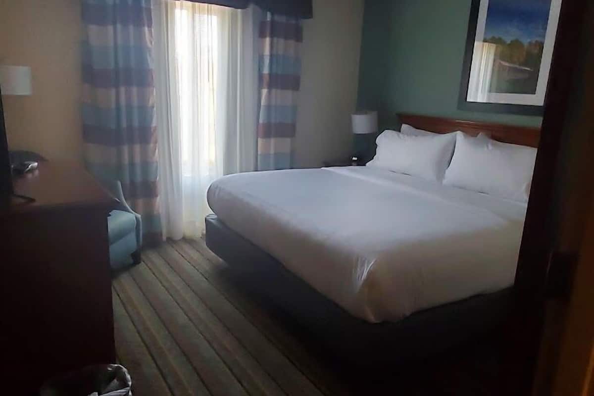 A hotel room with a large bed, a desk, a chair, a trash bin, and a window with curtains.