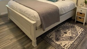 Una camera da letto con un letto, un comodino, una finestra con tende, un tappeto e un'opera d'arte a parete.