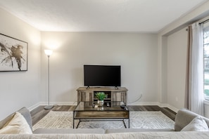 Un salon avec un téléviseur à écran plat posé sur un meuble en bois, une table basse en verre agrémentée d’une plante, un canapé et une lampe sur pied.