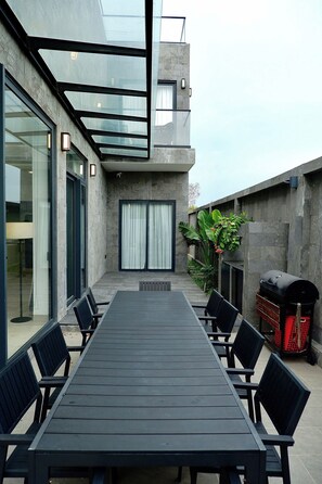 A modern outdoor dining area with a long table and chairs, a grill, and a view of the building's interior through glass walls. - Perolas Villas Resort - Ke Ga Bay (Tan Thanh)