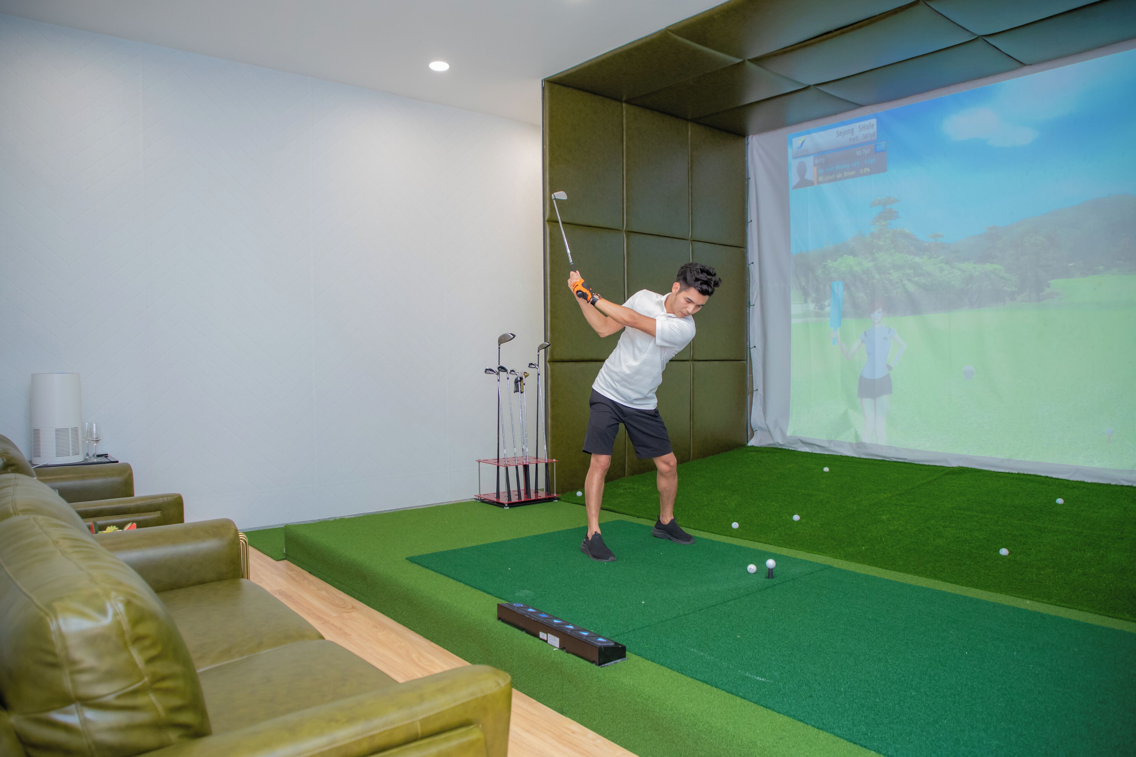 Un hombre practicando golf en una sala con una zona de césped verde, una pantalla de proyector mostrando un campo de golf y un área de descanso con sofás verdes.