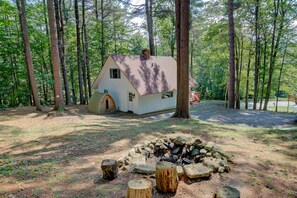 Una cabaña pequeña con chimenea, rodeada de árboles altos y un pozo de fuego al frente.