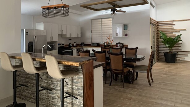Una cucina moderna con mobili bianchi, un'isola centrale con sgabelli da bar, una ventola a soffitto e una pianta in vaso.