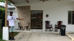 Un portico coperto con pavimento in cemento, soffitto in legno e parete in mattoni. Si vedono due sedie in legno e un tavolino. Sul lato destro è presente un cestino nero.