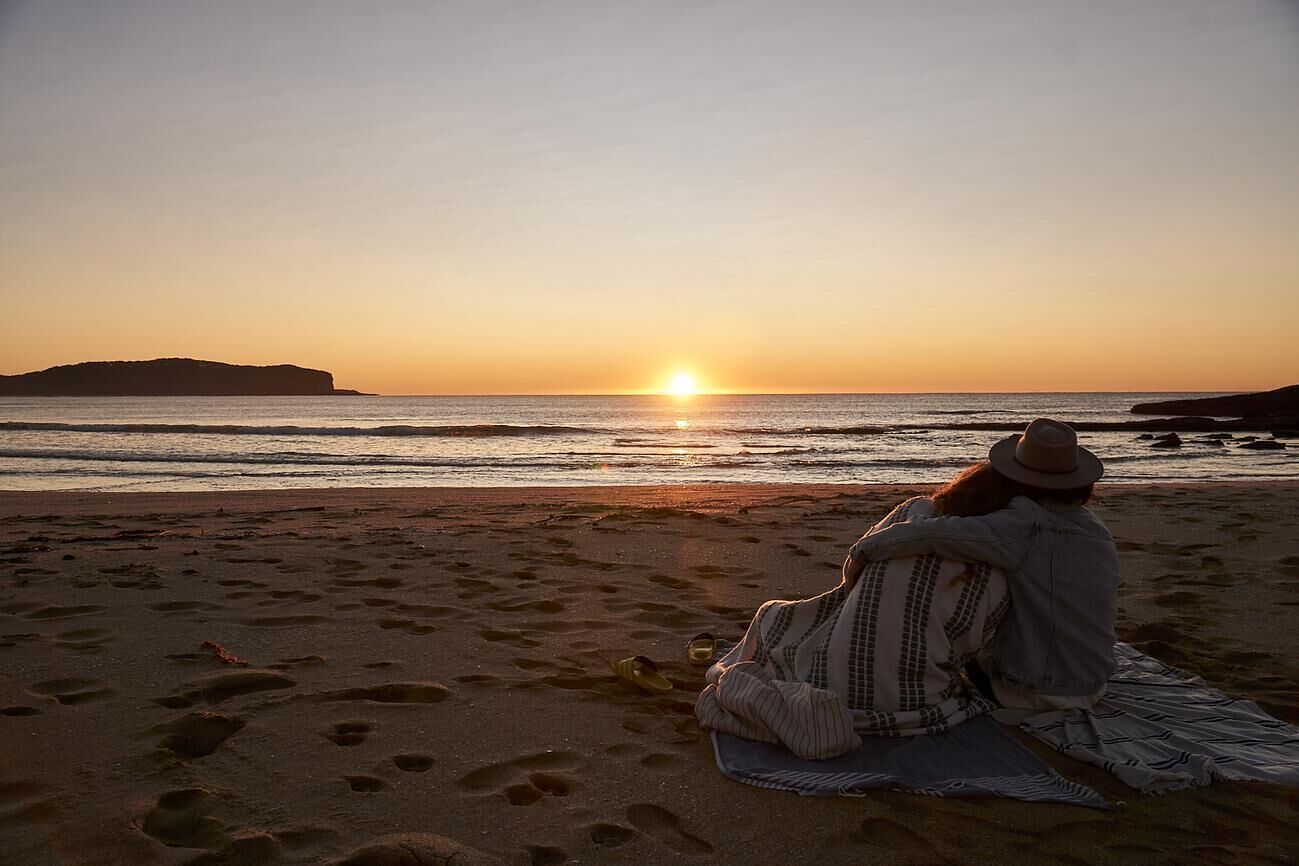Une vue du coucher de soleil sur une plage, avec des empreintes de pas dans le sable et une personne assise sur une couverture.