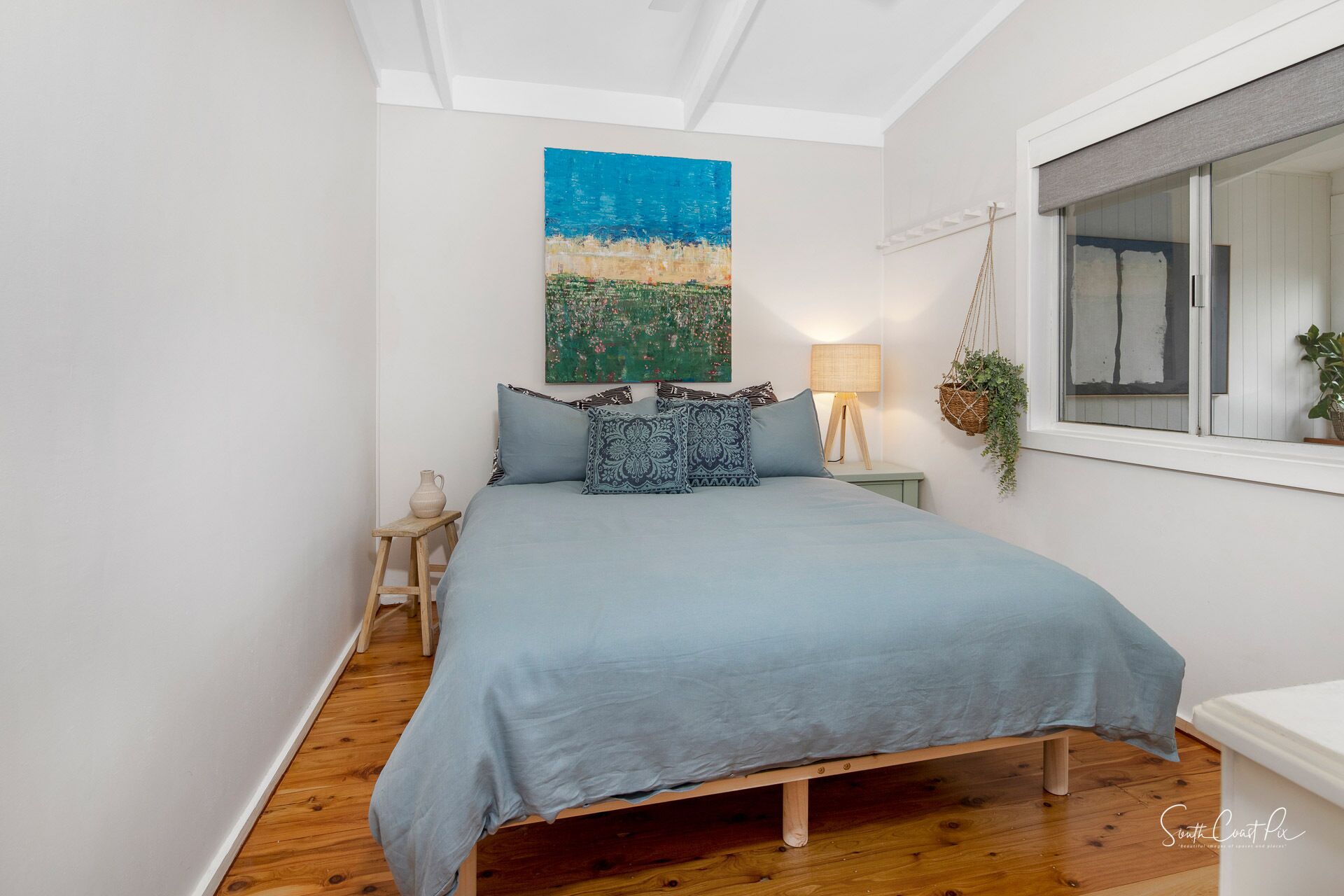 A bedroom with a bed, a hanging plant, a lamp, and a painting on the wall.