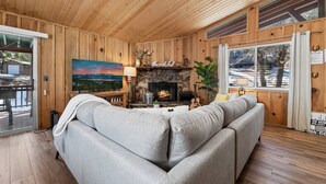 L’intérieur d’un chalet chaleureux, avec une cheminée en pierre, un canapé gris, des murs en bois et une grande fenêtre donnant sur un paysage enneigé.