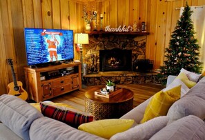 A cozy living room with a fireplace, a television displaying a Christmas program, a guitar, and a Christmas tree.