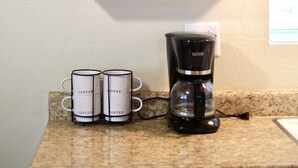 Un comptoir de cuisine avec une machine à café, deux tasses à café, un pot de sucre, une petite plante en pot et plusieurs verres à vin sur une étagère.