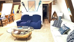 A cozy living room with a blue sofa, a glass coffee table, and a white couch with patterned pillows. There is a wooden dining table with chairs, a map on the wall, and a sign indicating the location.