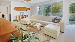 A modern living room with a large sofa, a coffee table, and a wooden dining table. There is a ceiling fan, a bookshelf, and a large window with a view of the outside. - Apartment 3 - The Cove (Noosa Heads)