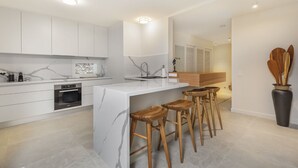 A modern kitchen with white cabinetry, a marble island, wooden stools, and a large vase with wooden utensils. - Apartment 3 - The Cove (Noosa Heads)