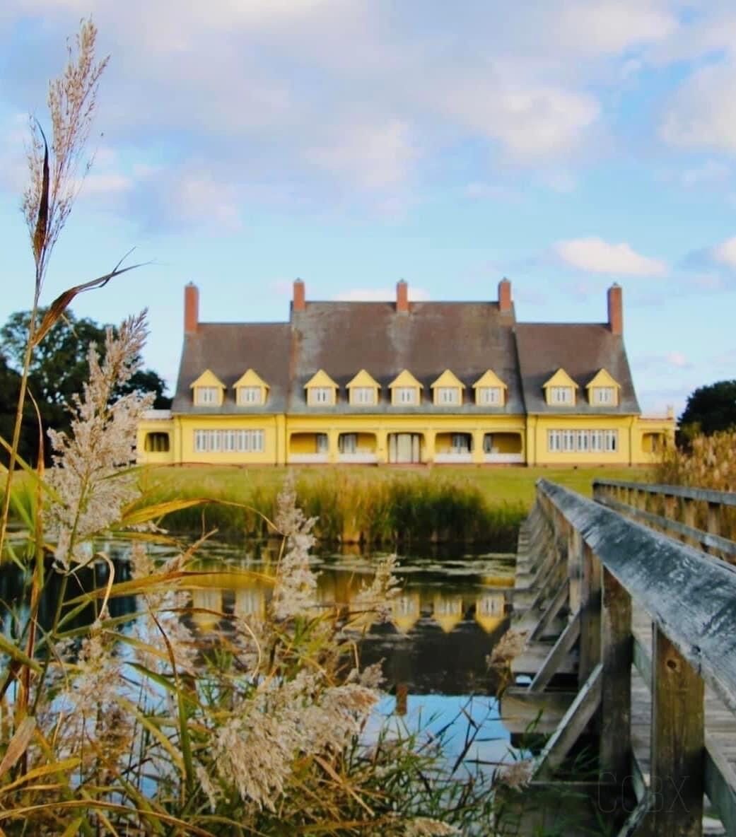A large yellow building with a symmetrical roof and multiple windows, situated by a calm body of water with a wooden bridge in the foreground.