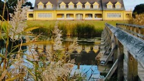 Een groot geel gebouw met een symmetrisch dak en veel ramen, gelegen aan een rustig water met een houten brug op de voorgrond.