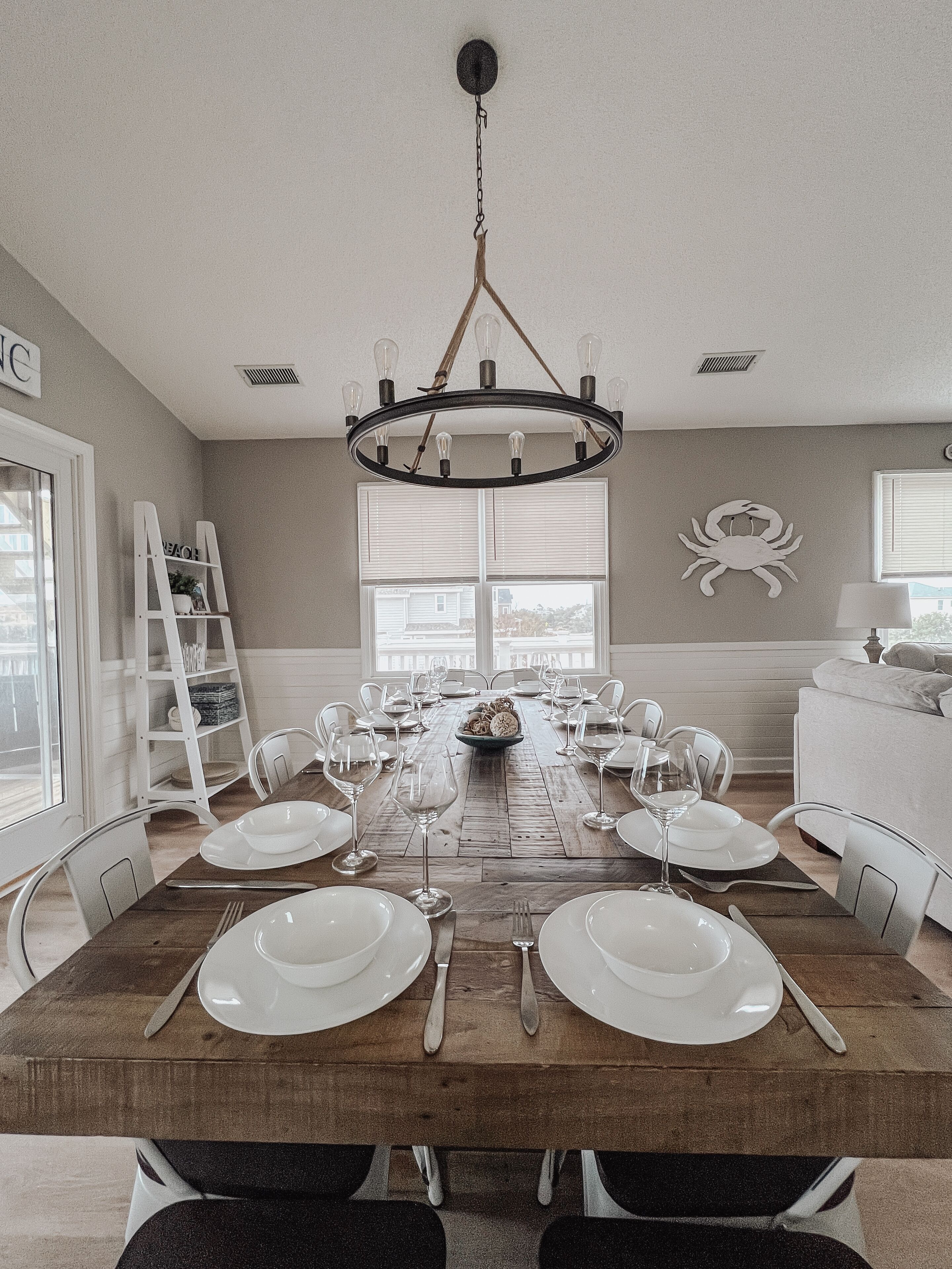 A dining room with a wooden table set for six, surrounded by white chairs, a chandelier, and a large window.