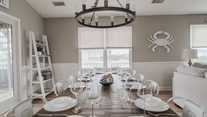Een eetkamer met een houten tafel gedekt voor zes personen, omringd door witte stoelen, een kroonluchter en een groot raam.