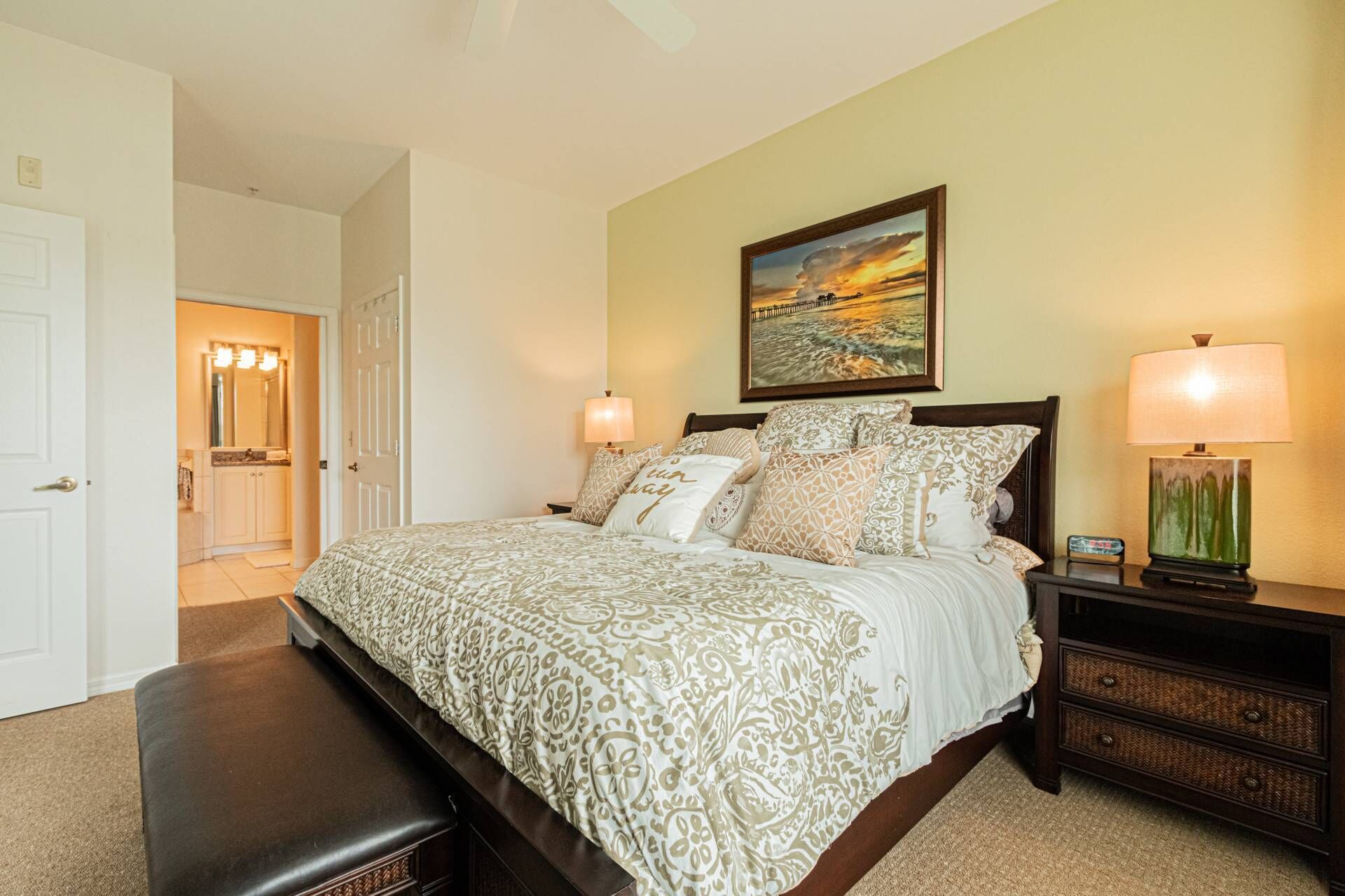 A bedroom with a large bed, two bedside lamps, a nightstand, a painting on the wall, and a door leading to another room.