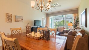 A dining area with a wooden table, chairs, and a chandelier. There is a television and a balcony with a view of the outdoors. - GreenLinks/Updated Lely Condo; w/Office space-Lake Views-Resort Amenities (Naples)