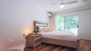 Une chambre avec un lit en bois, une table de chevet, une chaise, un ventilateur de plafond et une fenêtre donnant sur un paysage verdoyant.