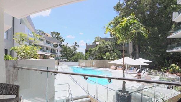 Un hôtel moderne doté d’une balustrade en verre donnant sur une piscine, le tout entouré d’une végétation luxuriante et d’autres bâtiments.