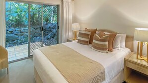 Un letto grande con coperta color beige e cuscini a righe, due abat-jour e vista su un muro in pietra e vegetazione attraverso una porta in vetro.