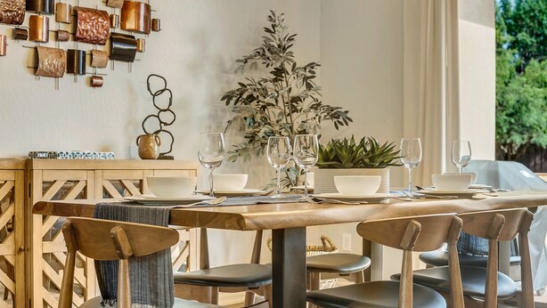 A dining area with a wooden table set for two, surrounded by chairs. There is a potted plant on the table, and a wall-mounted decorative piece above it. - Experience luxury in this 3-bedroom home with stunning red rock views in Sedona. (Sedona)