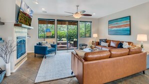 A spacious living room with a large brown leather sofa, a blue armchair, and a dining area with a table and chairs. There is a fireplace with a blue accent wall, a ceiling fan, and large windows offering a view of the outdoors. - Experience luxury in this 3-bedroom home with stunning red rock views in Sedona. (Sedona)