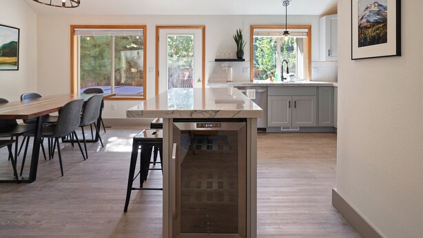A modern kitchen with a central island, large windows, and a dining area with chairs. - Quail Covey - Hot Tub, AC, Beautiful, Fully Fenced Yard, Close to Everything! (Bend)