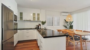 A modern kitchen with white cabinetry, a central island, and a dining area with wooden chairs and a table. - Apartment 7 - The Cove (Noosa)