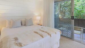 A bedroom with a large bed, a nightstand, a lamp, and a view of a balcony with a chair and plants. - Apartment 7 - The Cove (Noosa)