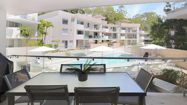 A balcony with a glass railing overlooking a swimming pool and a multi-story building with balconies. - Apartment 7 - The Cove (Noosa)