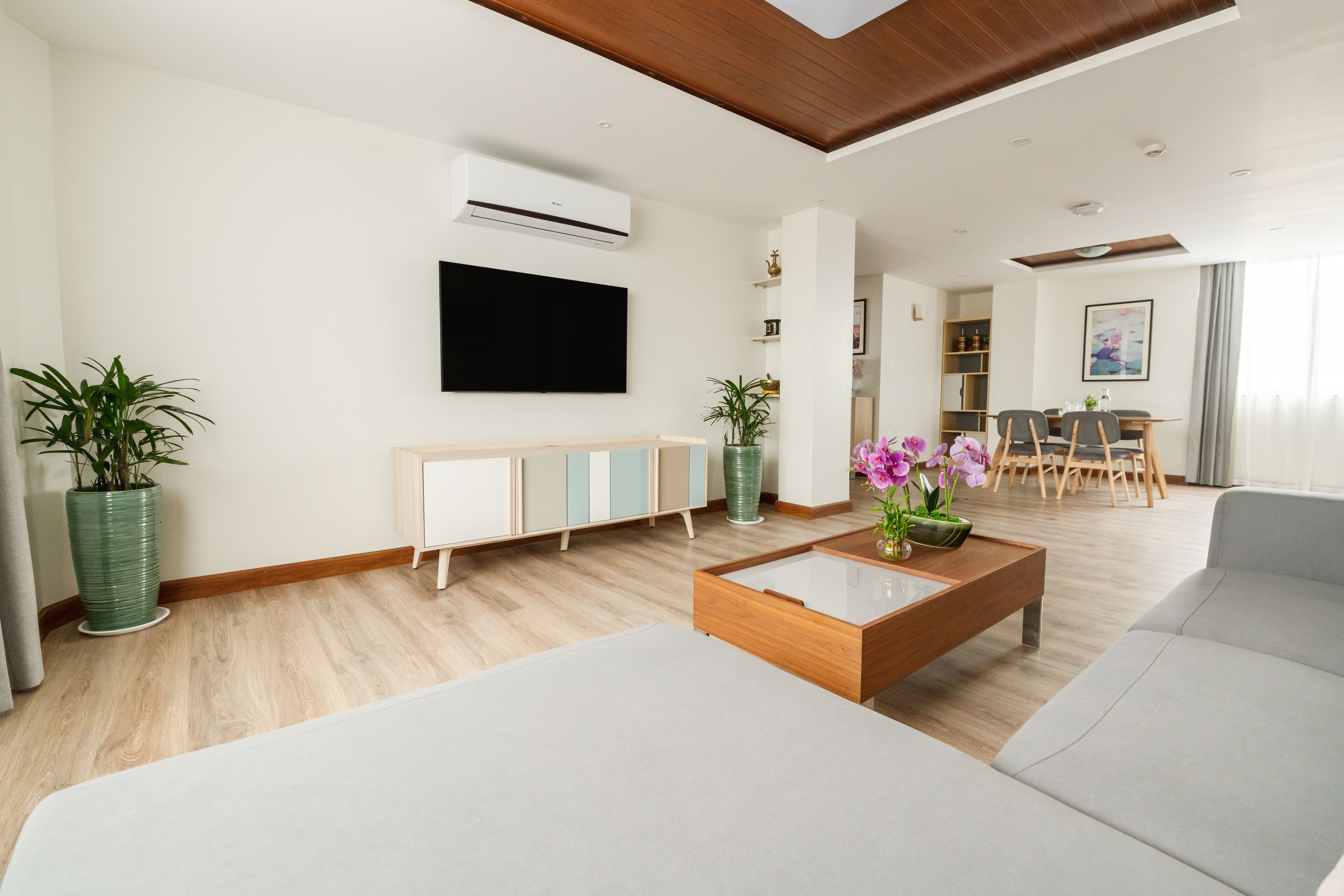 A modern living room with a flat-screen TV, a wooden coffee table, and a grey sofa. There is a dining area in the background with a table and chairs.