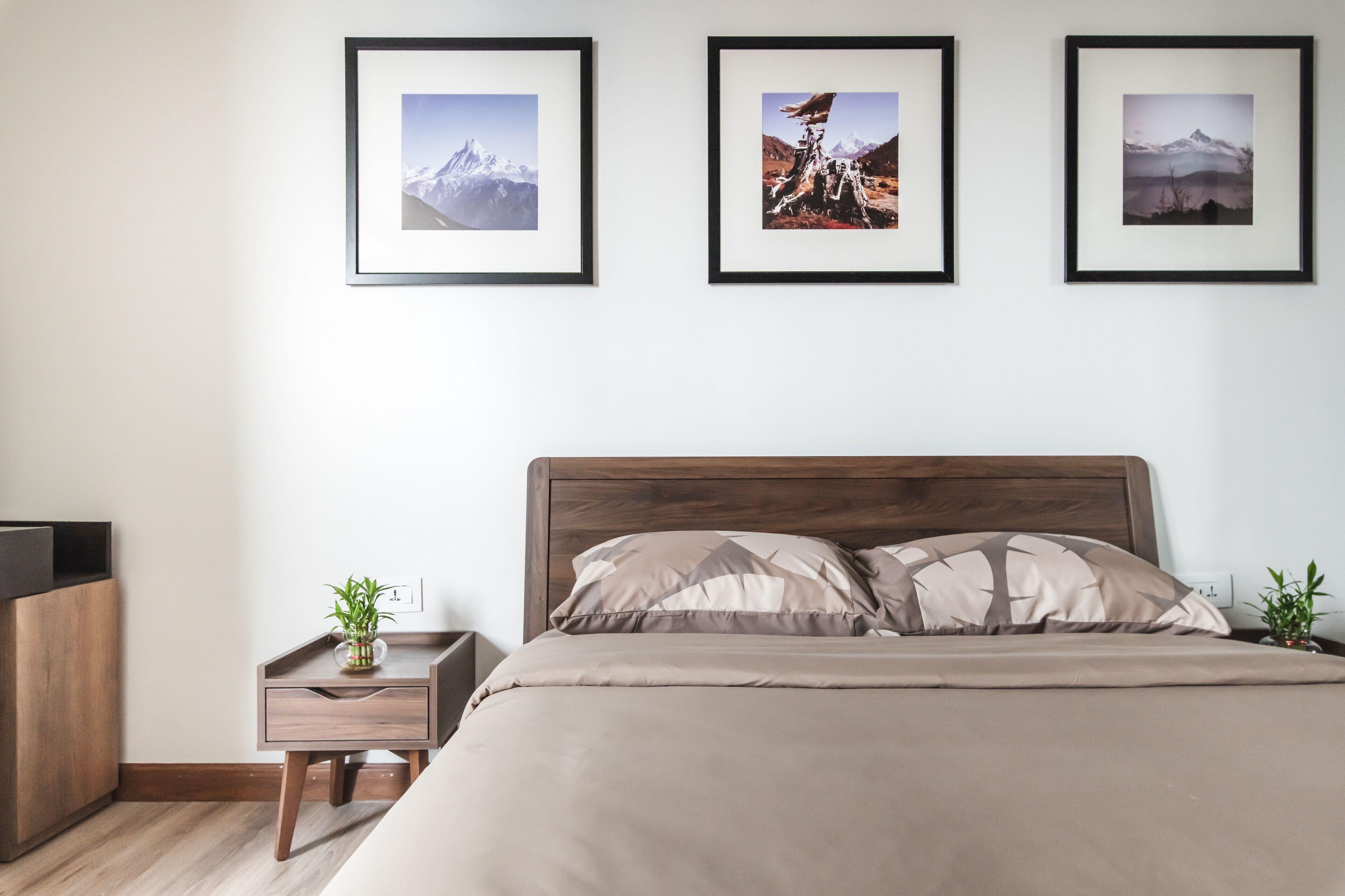 Une chambre à coucher avec une tête de lit en bois, un lit avec une literie beige, une table de chevet avec une plante et trois tableaux encadrés accrochés au mur.