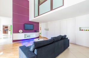 Ein modernes Wohnzimmer mit einem großen grauen Sofa, einem Flachbildfernseher an einer violetten Akzentwand und einer weißen Kommode mit Glasplatte.