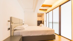 A modern bedroom with a large bed, a bedside table, a wardrobe, and a view of the outside through large windows.