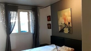 Ein Schlafzimmer mit Bett, Fenster mit Vorhängen, Deckenleuchte und einem Bild an der Wand.