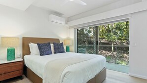 A bedroom with a large bed, a nightstand, a lamp, and a view of a garden through a sliding glass door. - Penthouse 9 - The Cove (Noosa Heads)