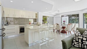 A modern kitchen with white cabinetry, a central island, and a dining area with white chairs. - Penthouse 9 - The Cove (Noosa Heads)
