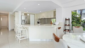 A modern kitchen with white cabinetry, a central island with a sink, and a dining area with a round table and chairs. - Penthouse 9 - The Cove (Noosa Heads)
