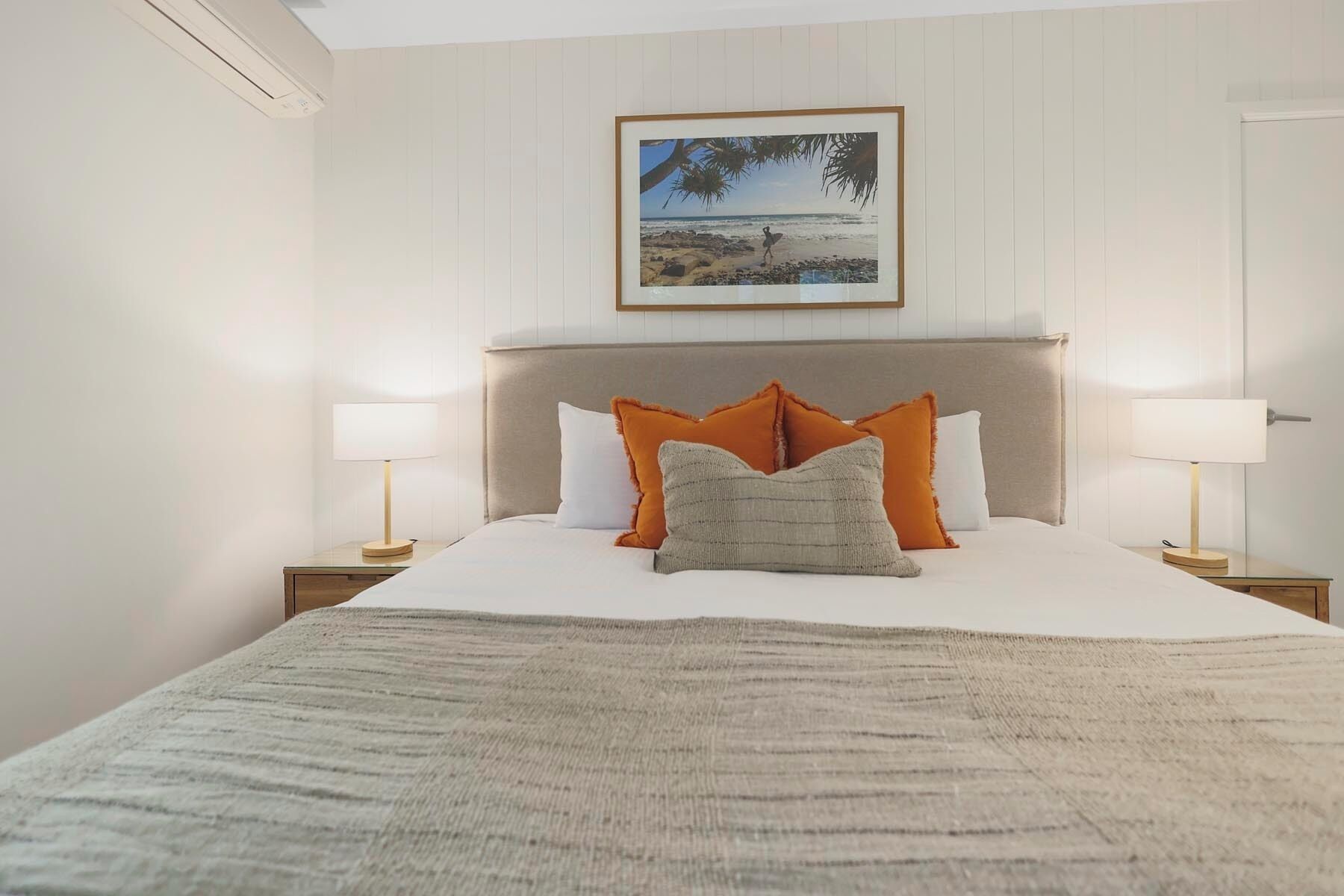 Ein ordentlich bezogenes Bett mit weißer Decke und drei Kissen, zwei orange und eines grau, in einem Zimmer mit weißen Wänden und einem über dem Bett hängenden Bild einer Strandansicht.